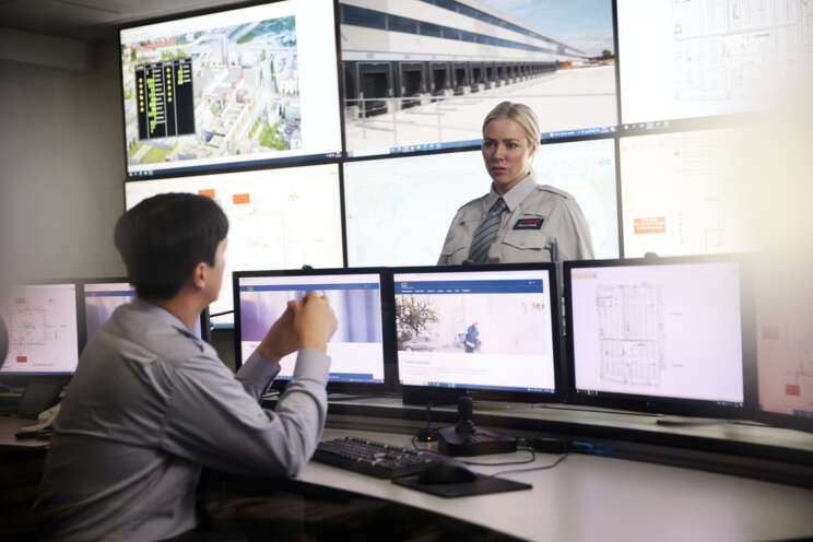 A Securitas security professional is talking with another Securitas security professional in a room with many monitors.